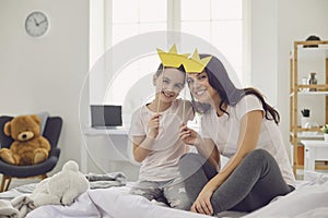 Happy mother`s day. Mother and daughter play princess with cardboard crowns on their heads while sitting on the bed.