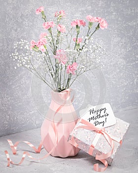 Happy mother's day note on gift box with ribbon, pink carnation, white gypsophila in vase on grey.