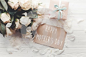 Happy Mother`s Day text sign on craft greeting card and white roses bouquet, gift box on wooden background, flat lay. Mothers day