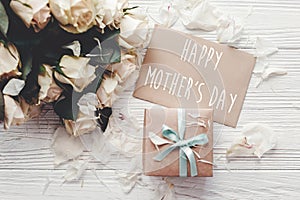 Happy Mother`s Day text sign on craft greeting card and white roses bouquet, gift box on wooden background, flat lay. Mothers day