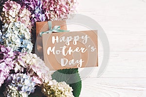 Happy Mother`s Day text sign on paper greeting card with gift box and beautiful hydrangea flowers on rustic white wooden table.