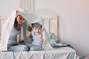happy mother and son playing and hiding under blanket in bed in cozy weekend morning.