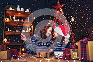 Holidays, family and people concept. Happy mother and little girl in santa helper hat with sparklers in hands, gift