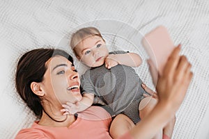 Happy mother taking selfie with her cute infant baby at home, lying on bed and capturing photo on cellphone