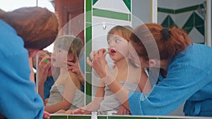 Happy mother is teaching her child to brush teeth patiently at home near mirror