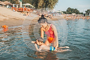 Happy mother teaching her little son swimming in the water, on the beach