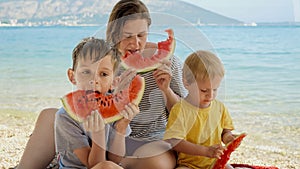 Happy mother with two boys sitting on the sea beach and enjoying eating sweet watermelon. Concept of family vacation