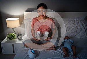 happy mother using breast pump machine to pumping milk with her newborn baby on bed at night