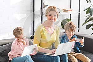 happy mother using laptop and smiling at camera while kids using digital devices