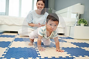 happy mother watching her infant baby crawlinf on play mat or jigsa floor in bedroom