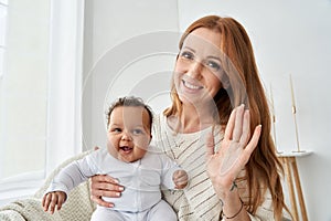 Happy mother waving hand holding funny baby daughter looking at camera at home.