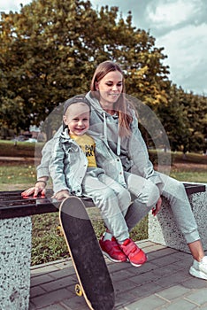 Happy mother woman with little boy son 4-5 years old, sitting on bench in summer autumn city park. Skateboard, casual