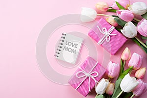 Happy mothers day concept. Top view of pink tulip flowers, gift box and note book with LOVE MOM text on pink pastel background.