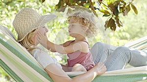 Happy mothers day, smiling mom playing with her blue eyed little girl daughter child with curly blonde hair, lying on hammock