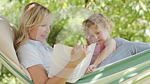 Happy mothers day, smiling mom reading story from the book to her blue eyed little girl daughter child with curly blonde hair,