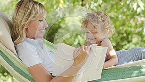 Happy mothers day, smiling mom reading story from the book to her blue eyed little girl daughter child with curly blonde hair,