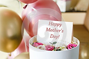 Happy Mothers Day text on gift card in flower box near festive rose and gold balloons. Greeting card for Mom. Flower delivery,