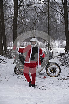 Happy motorcyclist in a Santa Claus suit shows likes. Winter forest with falling snow. Touring motorcycle in the background. The