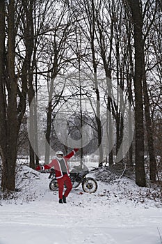 Happy motorcyclist in a Santa Claus suit shows likes. Winter forest with falling snow. Touring motorcycle in the background. The