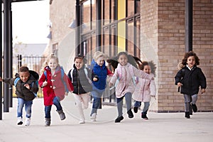 A happy multi-ethnic group of young school kids wearing coats and carrying schoolbags running in a walkway with their classmates o