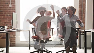 Happy multicultural office workers laughing having fun riding on chairs