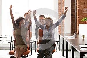 Happy multicultural office workers laughing having fun riding on chairs