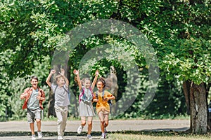 Happy multicultural schoolkids gesturing with raised hands while running in park