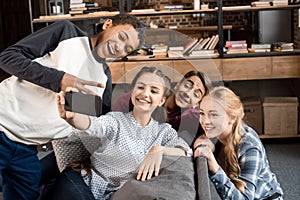 Happy multicultural teenagers group taking selfie on smartphone and sitting on sofa at home