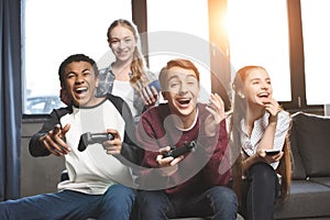 Happy multicultural teenagers playing video games with joysticks at home