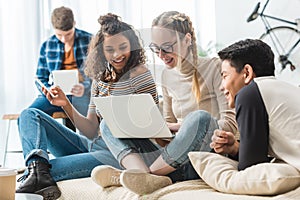 happy multicultural teens looking at laptop and sitting