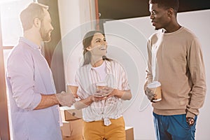 happy multiethnic coworkers holding paper cups and talking in new office