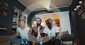 Happy multiethnic male friends shout and cheer watching sports game together at home on TV using projector slow motion.