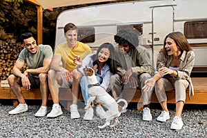 Happy multiethnic young friends sitting near RV, playing with pet dog, enjoying autumn vacation on campsite