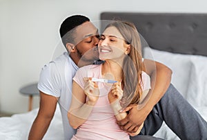 Happy multinational couple celebrating positive result of pregnancy test on bed at home