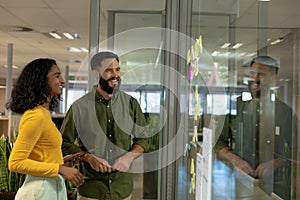 Happy multiracial colleagues looking at strategy on glass wall while discussing at modern workplace