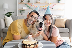 Happy multiracial couple celebrating their dog's birthday with festive cake, wearing party hats at home