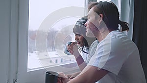 Happy multiracial couple drinking hot beverage while looking through the window at first snow