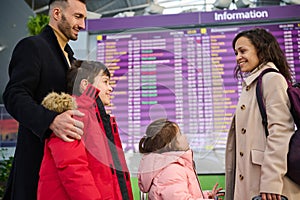 Happy multiracial family traveling together standing in front of the check-in board in the airport departure hall. Beautiful woman