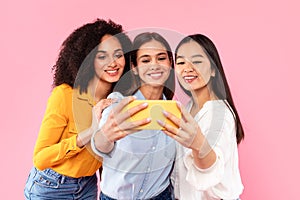 Happy multiracial female friends making selfie on cellphone, taking photos for social media over pink background