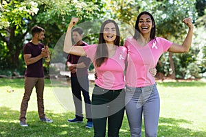 Happy multiracial female friends wearing breast cancer awareness ribbons flexing muscles in park