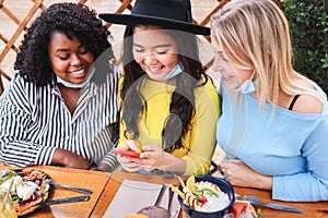 Happy multiracial friends using mobile phone outdoors at brunch restaurant with mask under chins - Focus on asian woman