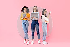 Happy multiracial ladies friends using different gadgets, using smartphone, laptop and digital tablet, pink background