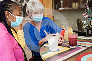 Happy multiracial senior woman using mobile phone at bar while wearing safety masks - Focus on right female