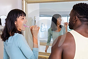 Happy multiracial young couple brushing teeth together in front of mirror at home