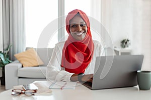 Happy muslim african american woman sitting at workplace and smiling at camera while working on laptop at home office