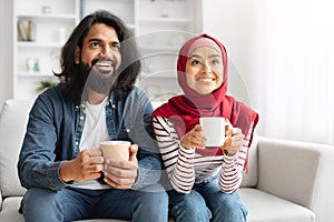 Happy Muslim Couple Drinking Coffee And Relaxing On Couch In Living Room