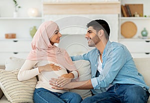 Happy muslim couple expecting baby, resting on couch at home and smiling, man touching his pregnant wife's belly