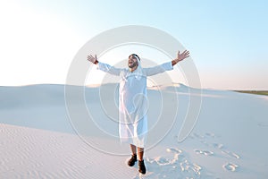 Happy Muslim man looks through banknotes and beside himself with joy, standing in center of sandy desert on warm, hot