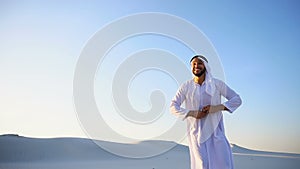 Happy Muslim man looks through banknotes and beside himself with joy, standing in center of sandy desert on warm, hot