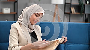 Happy Muslim woman in beige hijab reading paper book at home relaxing enjoy time for yourself